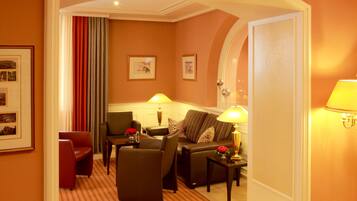 A hotel room with a seating area featuring a sofa, two armchairs, and a small table with a lamp. There are framed pictures on the wall and a door leading to another room.
