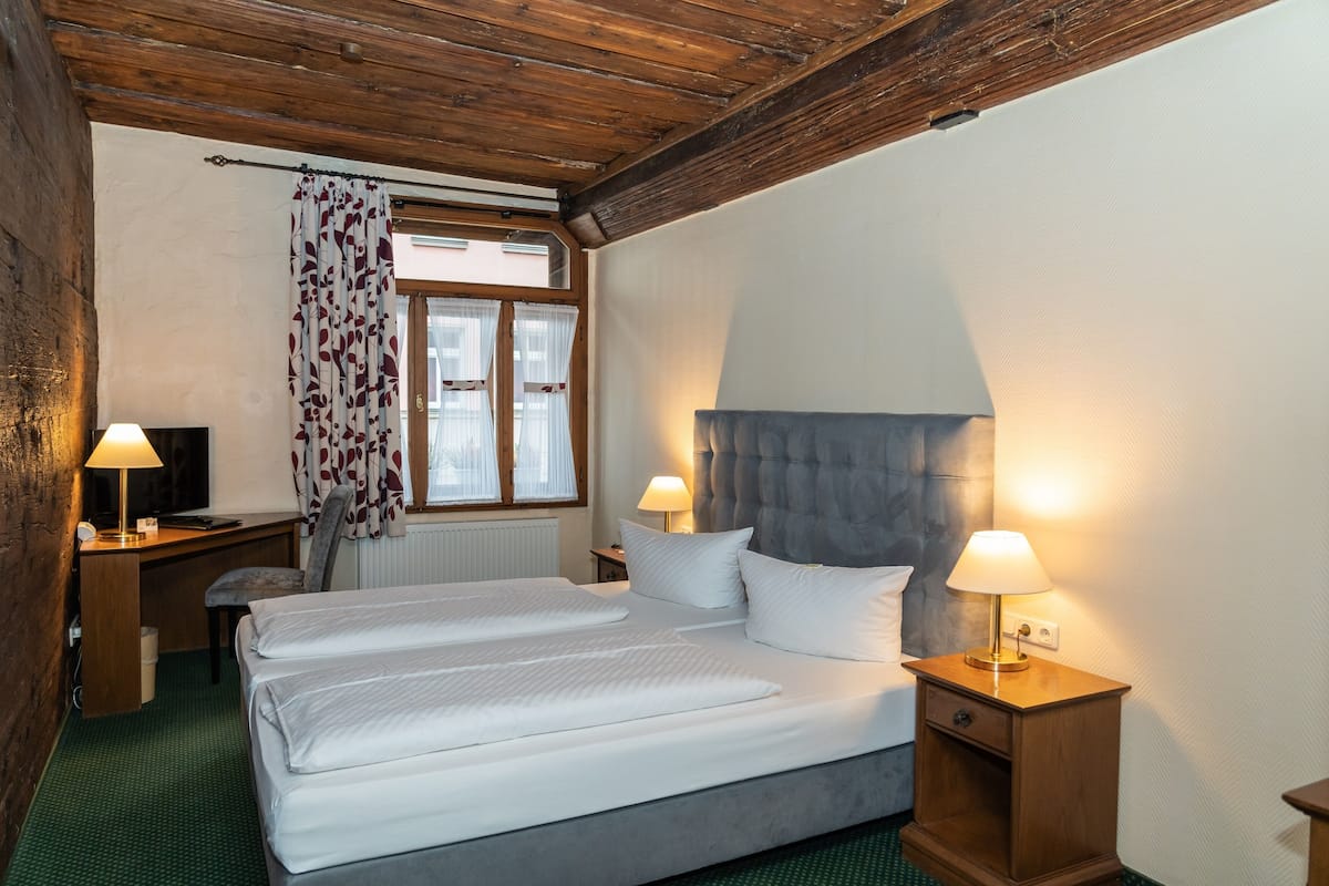 Una habitación de hotel con una cama doble, dos mesitas de noche con lámparas, un escritorio con silla, un televisor y una ventana con cortinas.