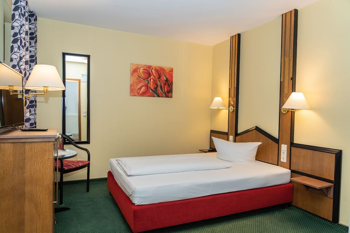 Una habitación de hotel con una cama individual, un escritorio con silla, un espejo y dos lámparas fijadas a la pared.