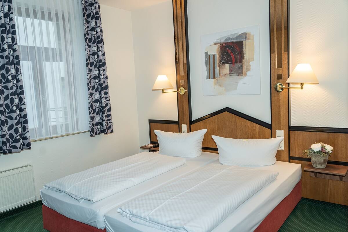 Habitación de hotel con cama doble, dos mesitas de noche, una lámpara de pared, una obra de arte enmarco y una ventana con cortinas.