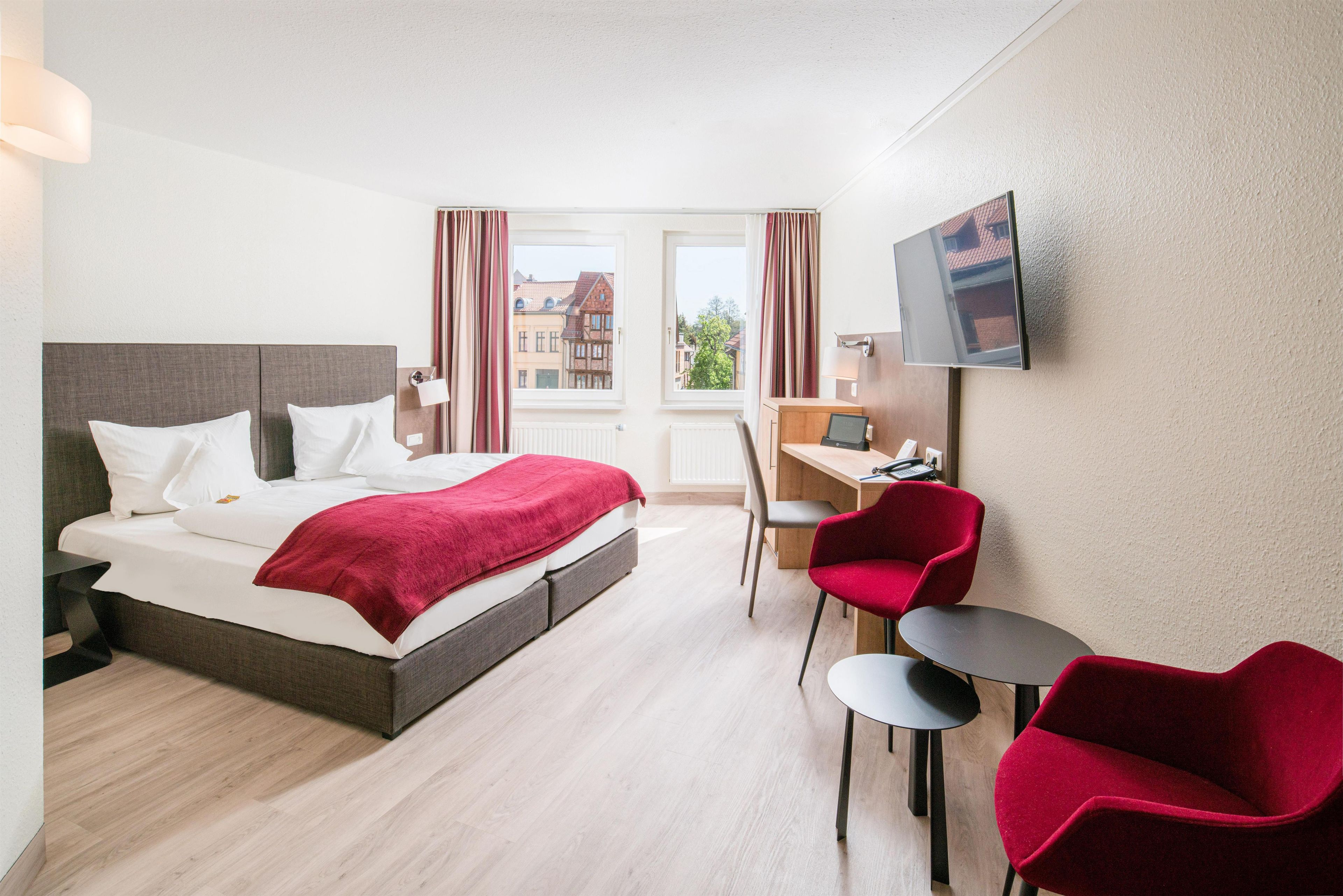 A hotel room with a bed, a desk, a chair, a TV, and a view of buildings outside.