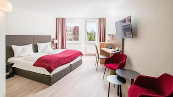 A hotel room with a bed, a desk, a chair, a TV, and a view of buildings outside.