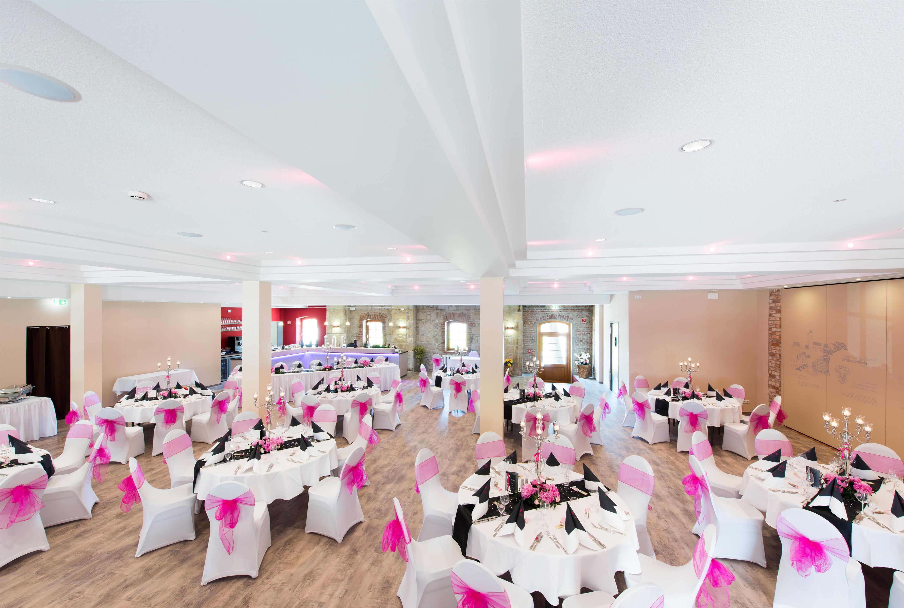 A spacious banquet hall with round tables and chairs, equipped with chandeliers and a bar area.