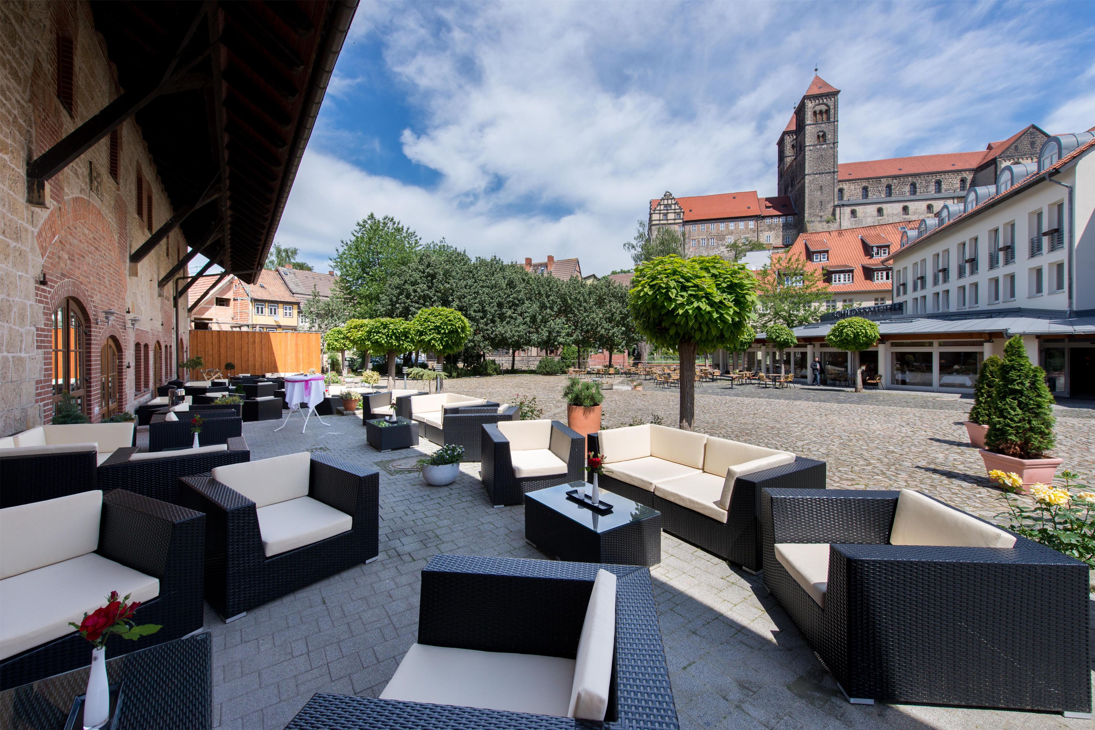 An outdoor seating area with wicker furniture and potted plants, set against a backdrop of historic buildings and a clear sky.