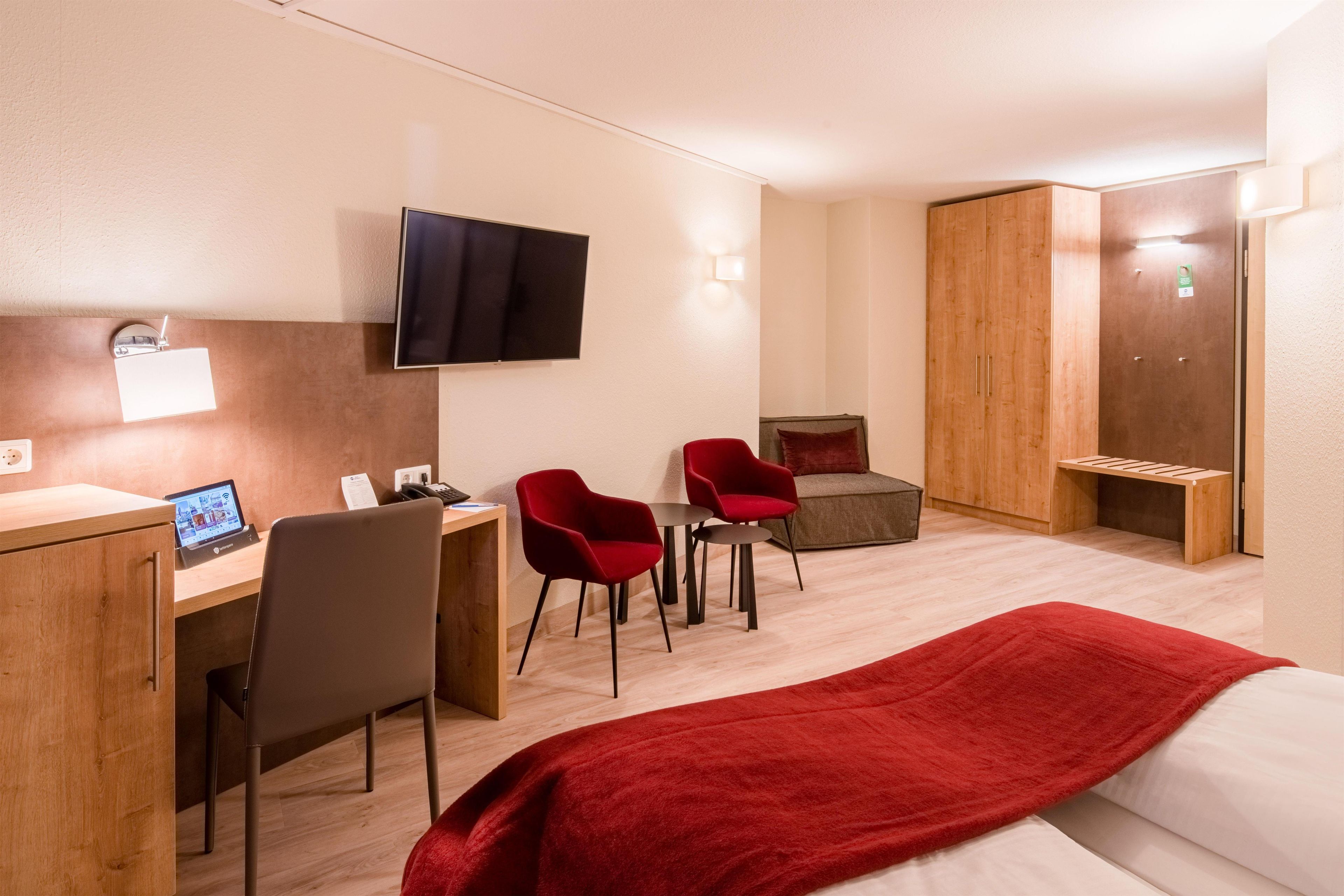 A hotel room with a bed, a desk with a computer, two chairs, a television, and a wardrobe.