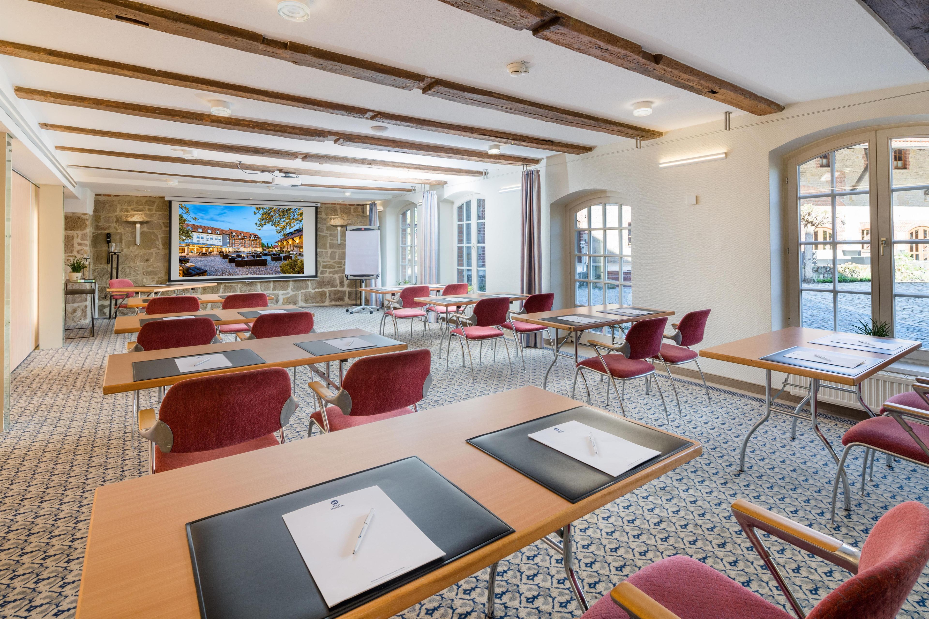 A conference room with tables, chairs, a projection screen, and a stone wall with a view of buildings.