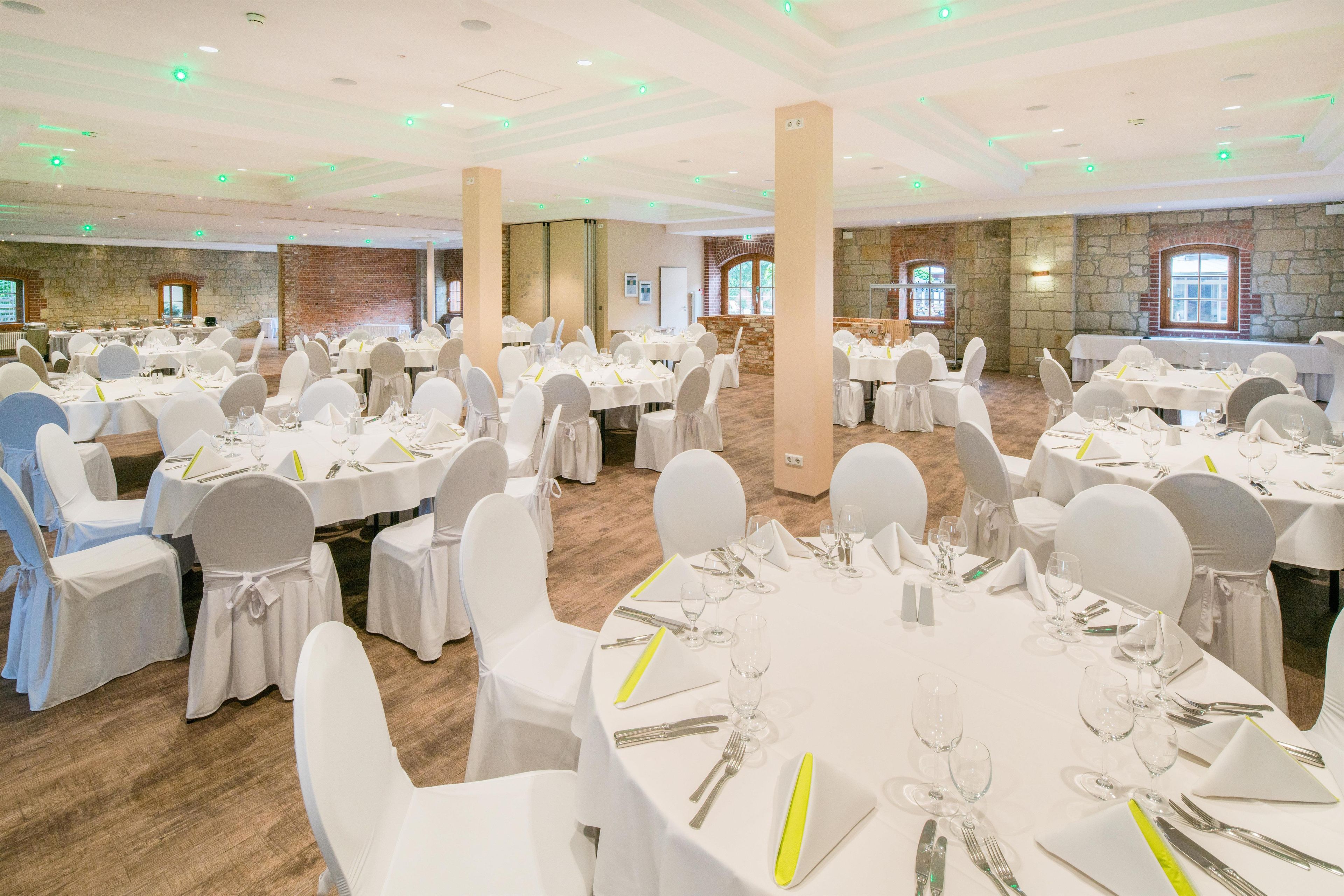 Un salón de banquetes con mesas redondas dispuestas para comer, sillas blancas y un fondo de pared de piedra.