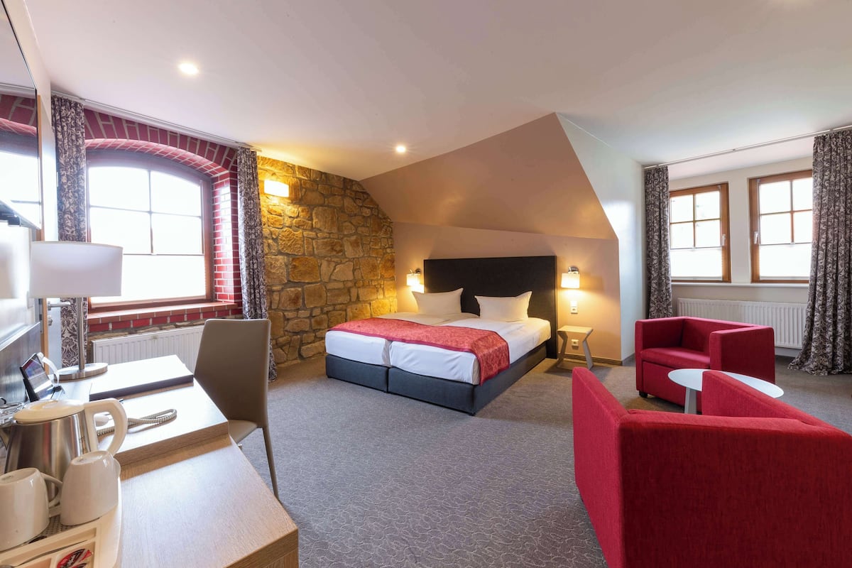 Une chambre d’hôtel avec un mur décoratif en pierre, un lit recouvert d’une couverture rouge, un bureau avec une machine à café et un fauteuil rouge.