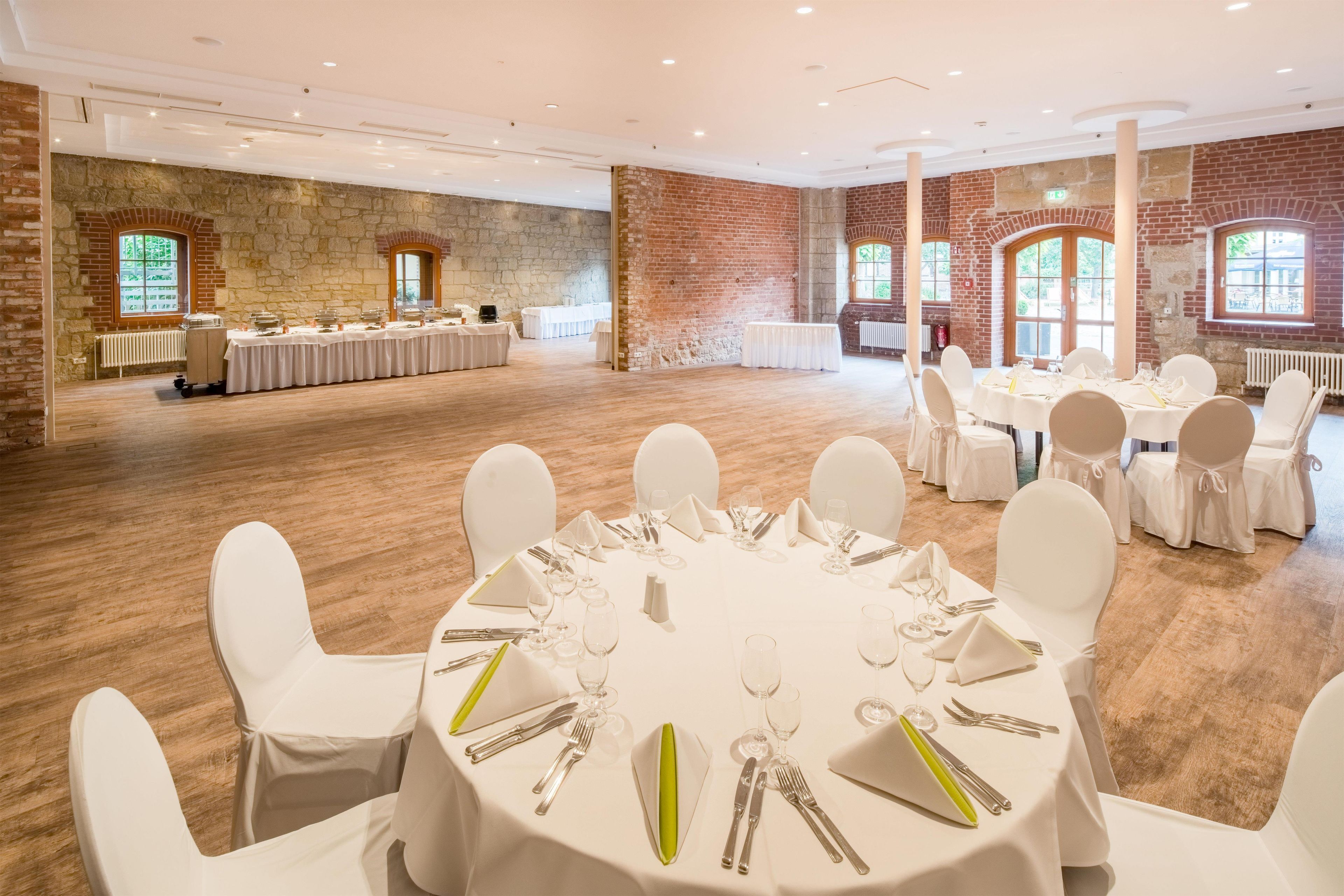 Un salón de banquetes con mesas redondas dispuestas para comer, sillas blancas y un fondo de pared de ladrillo.