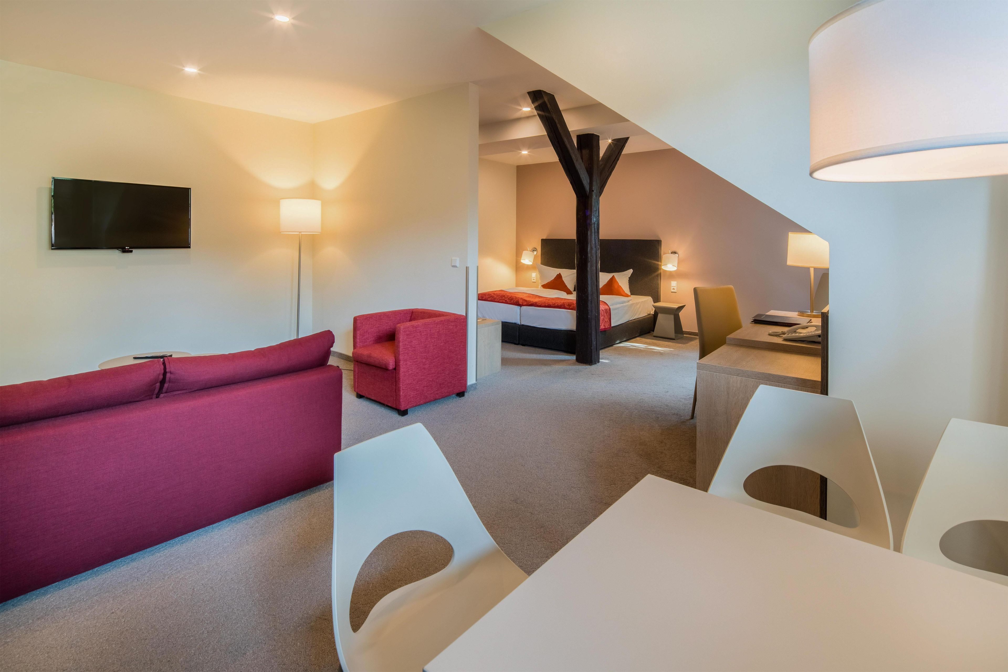 A modern hotel room with a flat-screen TV, a red sofa, a bed with white bedding, a desk with a chair, and a large window.