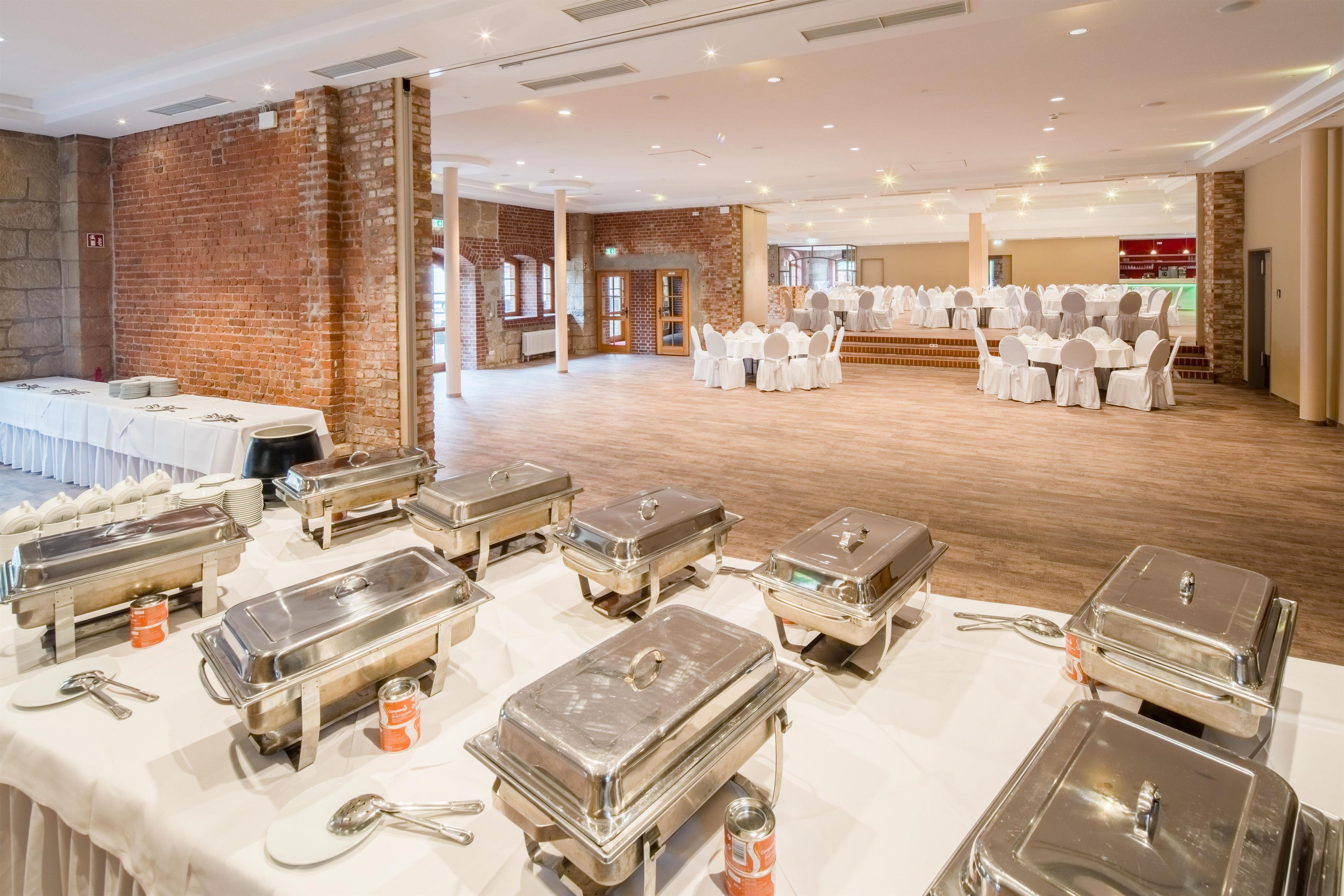 Un salón de banquetes con mesas preparadas para eventos, un bufé con varios recipientes de comida y una pared de ladrillo.