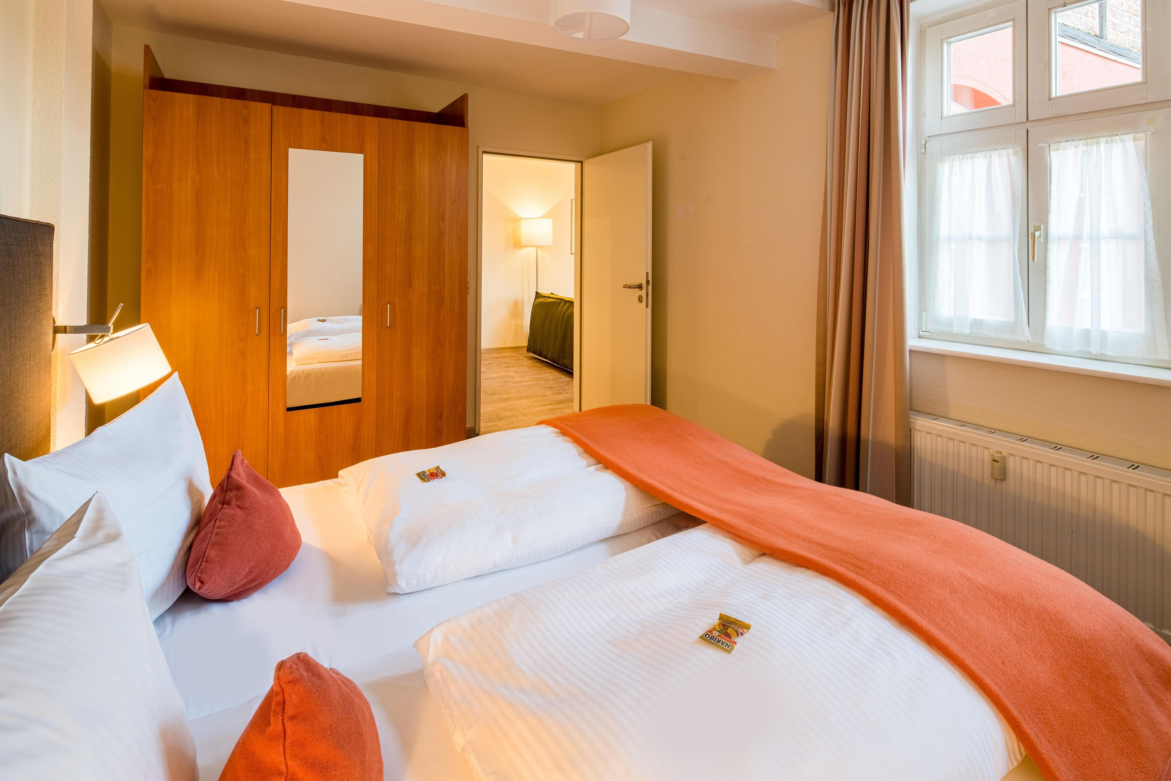 Quarto de hotel com cama, guarda-roupa, mesa de cabeceira, luminária e janela com cortinas.