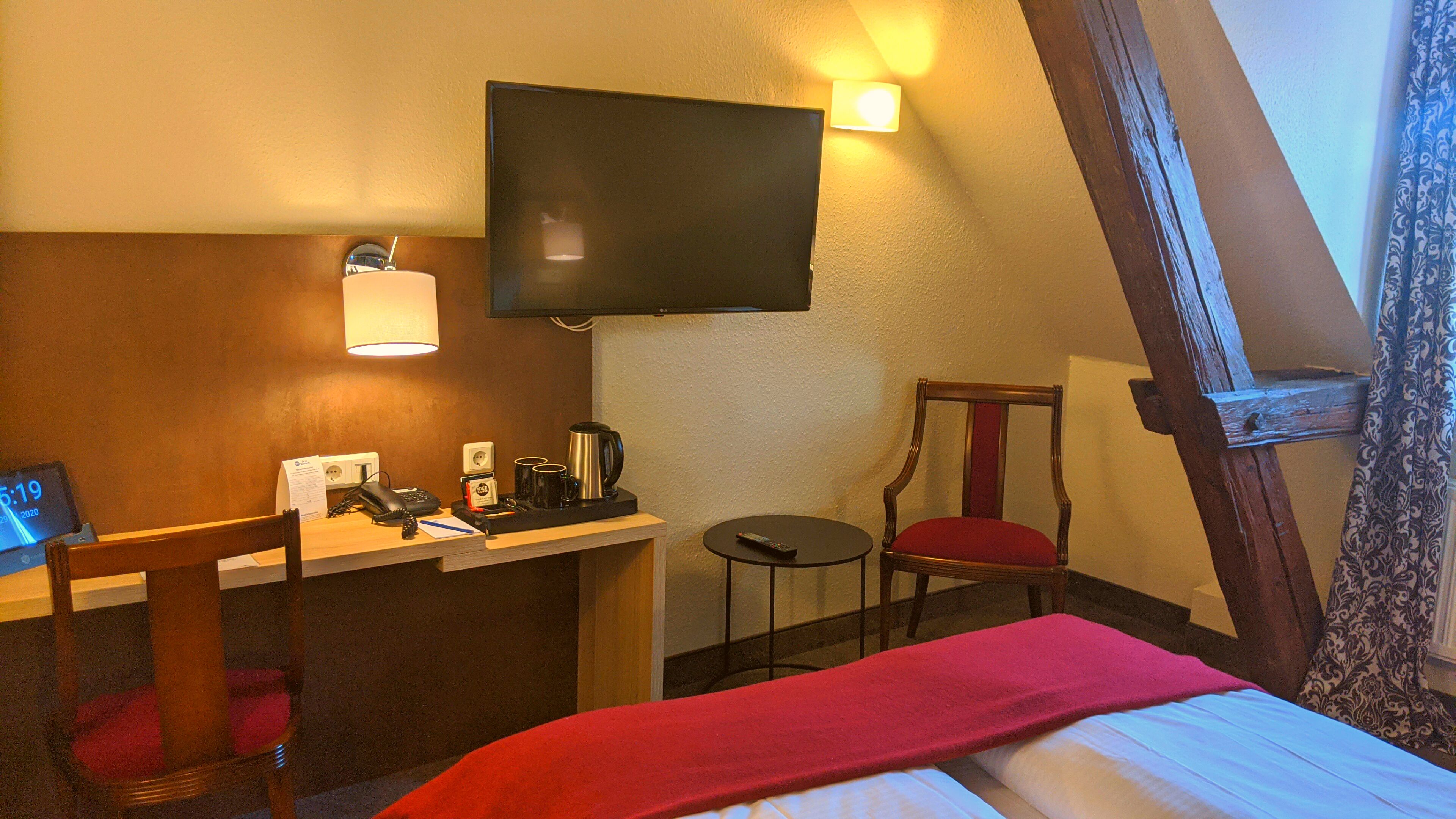 Een hotelkamer met een bed, een bureau met telefoon en koffiezetapparaat, een stoel, een aan de muur bevestigde televisie en een klein tafeltje.