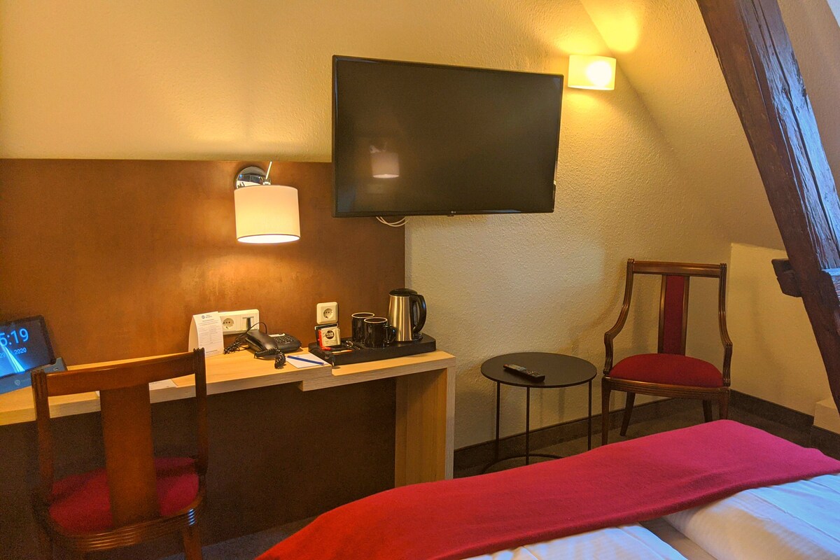 A hotel room with a bed, a desk with a telephone and a coffee maker, a chair, a television mounted on the wall, and a small table.