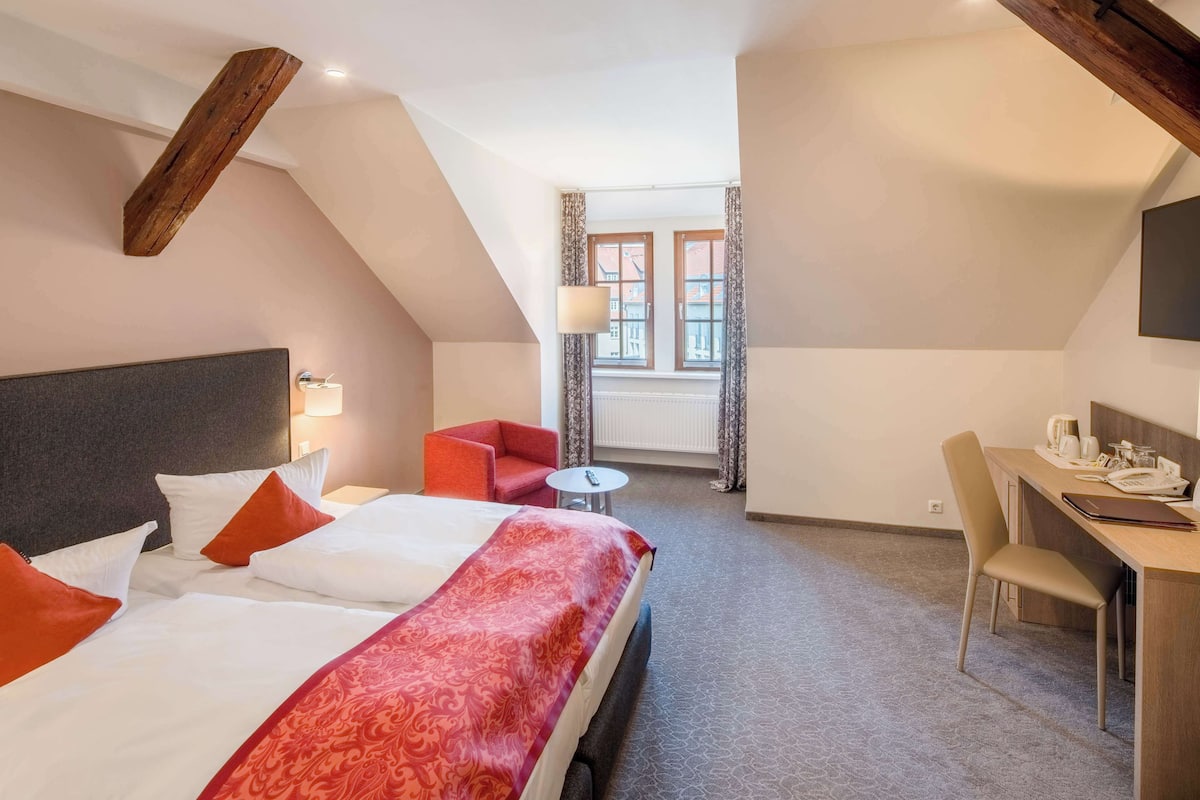 Une chambre d’hôtel avec un grand lit, un bureau, une chaise, une petite table et une fenêtre avec des rideaux.
