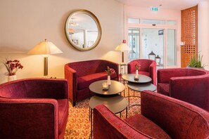 Un hall d’hôtel avec des sièges rembourrés de tissu rouge, deux tables rondes ornées de bougies, un grand miroir au mur et une moquette à motifs.