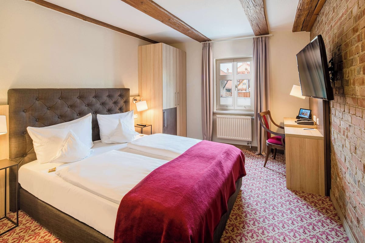 Une chambre d’hôtel avec un grand lit, un bureau avec une chaise, un téléviseur fixé au mur et une fenêtre avec des rideaux.