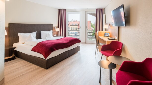 A hotel room with a bed, a desk, a chair, a TV, and a view of buildings through a door. - Best Western Hotel Schlossmuehle (Quedlinburg)