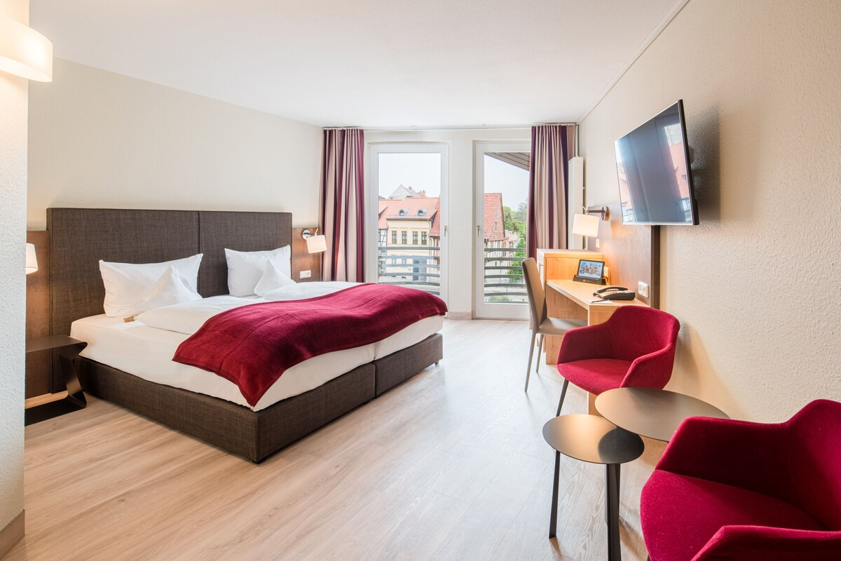 A hotel room with a bed, a desk, a chair, a TV, and a view of buildings through a door.