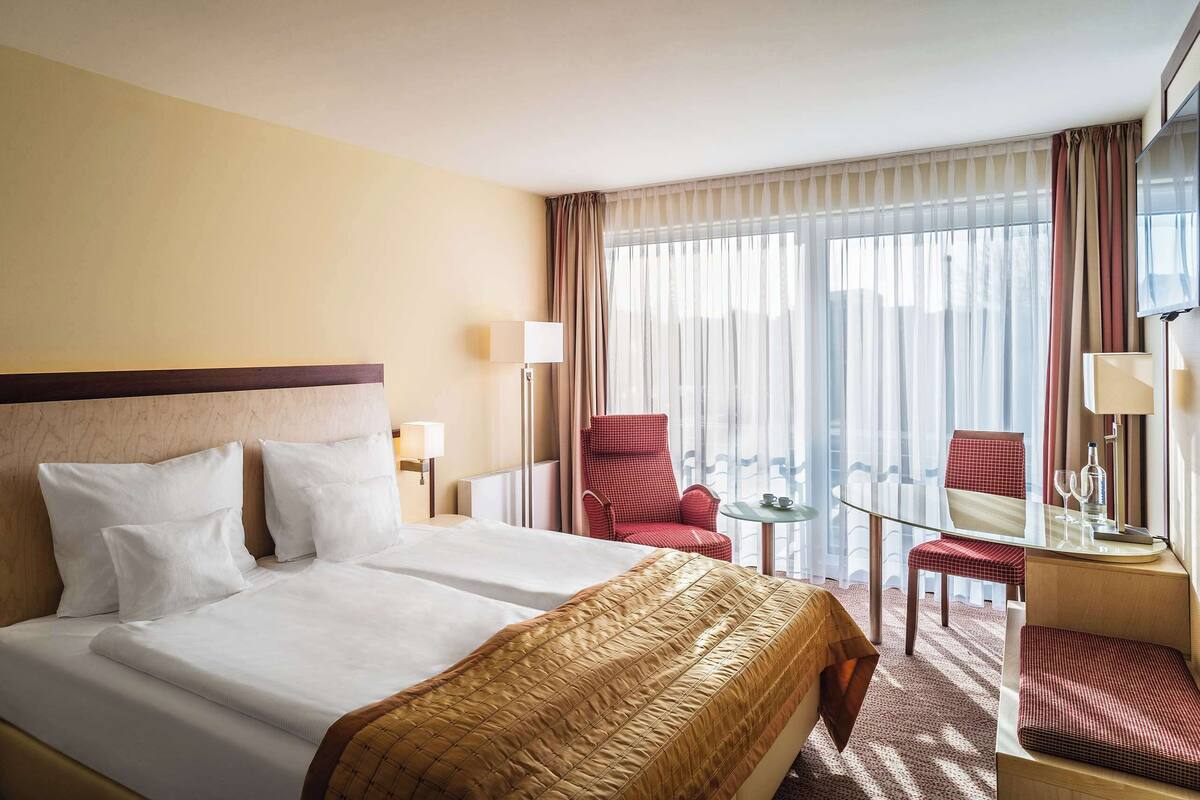 A hotel room with a large bed, two bedside tables, a desk with a chair, and a view of the outdoors through sheer curtains.
