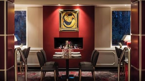 A dining area with a table set for two, a framed artwork above the table, and a view through the window.