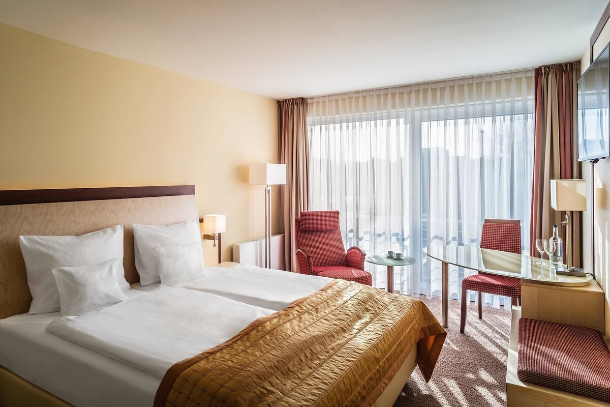 A hotel room with a large bed, a desk with a chair, a small table, and a view of the outdoors.