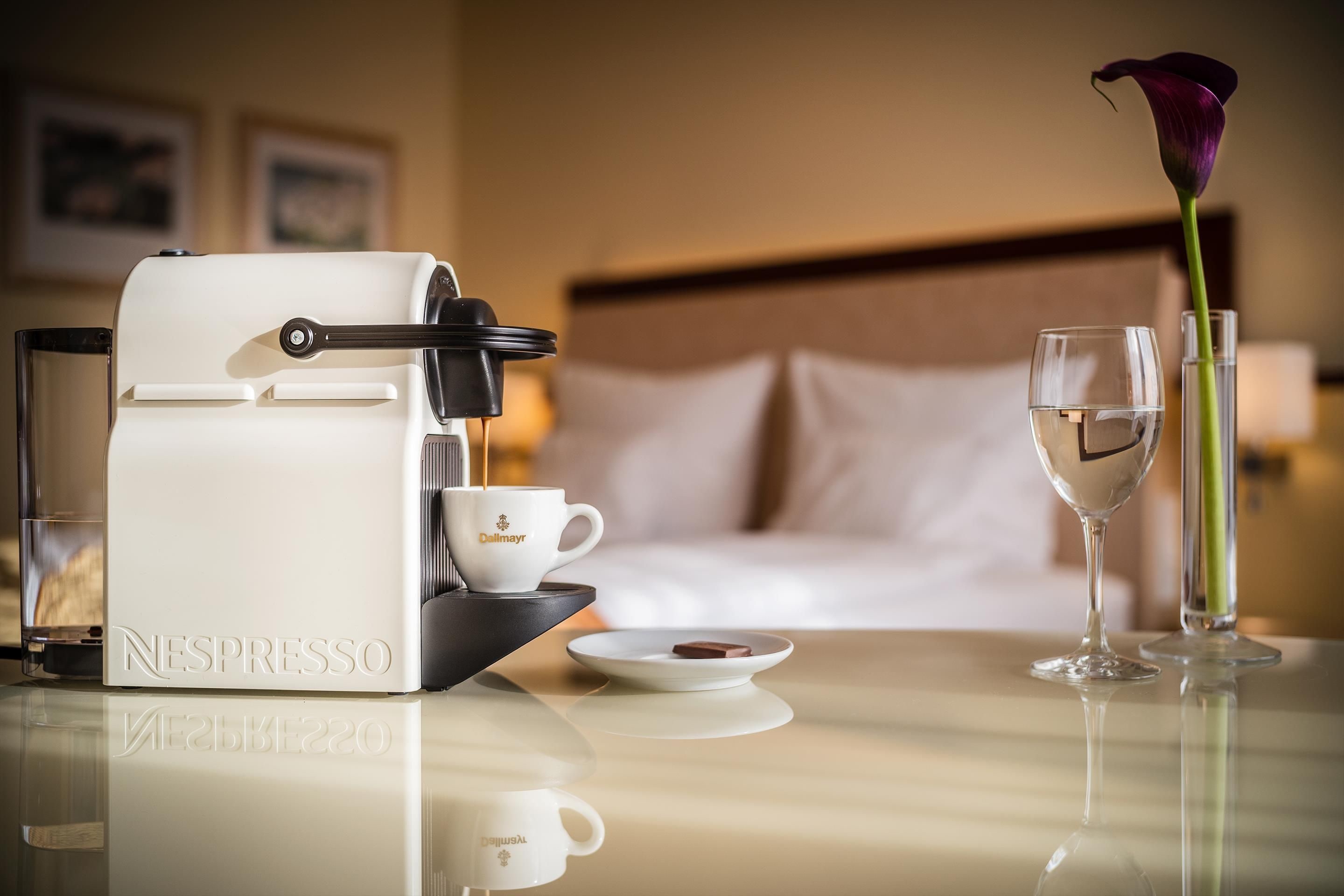 Una máquina de café Nespresso sobre una mesa con una taza, un vaso de agua y una flor.