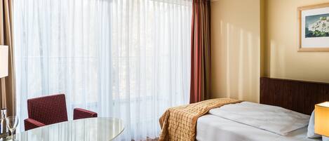 A hotel room with a bed, a table, two chairs, a window with curtains, and a painting on the wall.