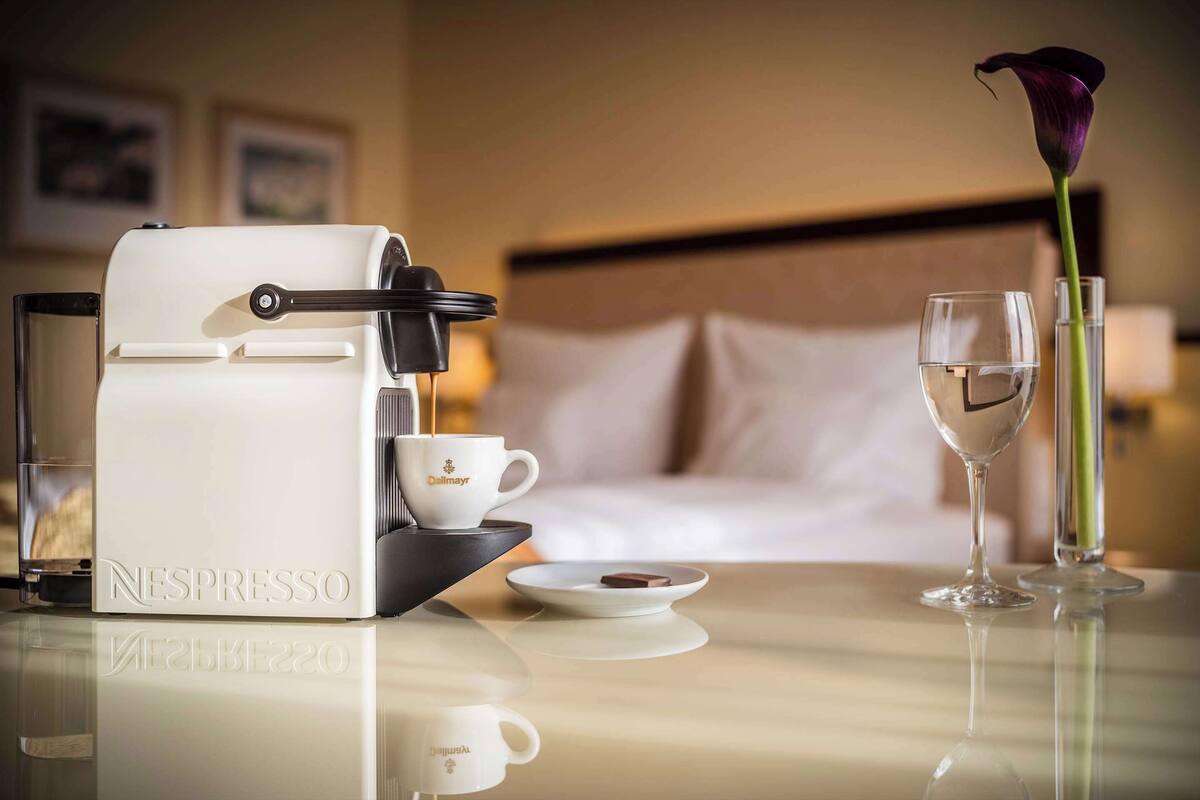 A Nespresso machine, a cup, a plate with a chocolate piece, a glass of water, and a vase with a flower on a hotel table.