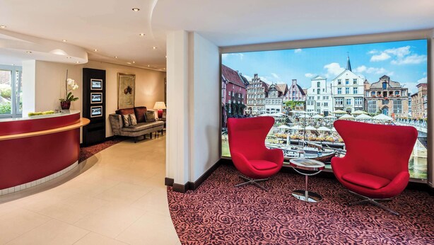 A hotel lobby with a reception desk, two red chairs, a patterned carpet, and a large window offering a view of a cityscape.