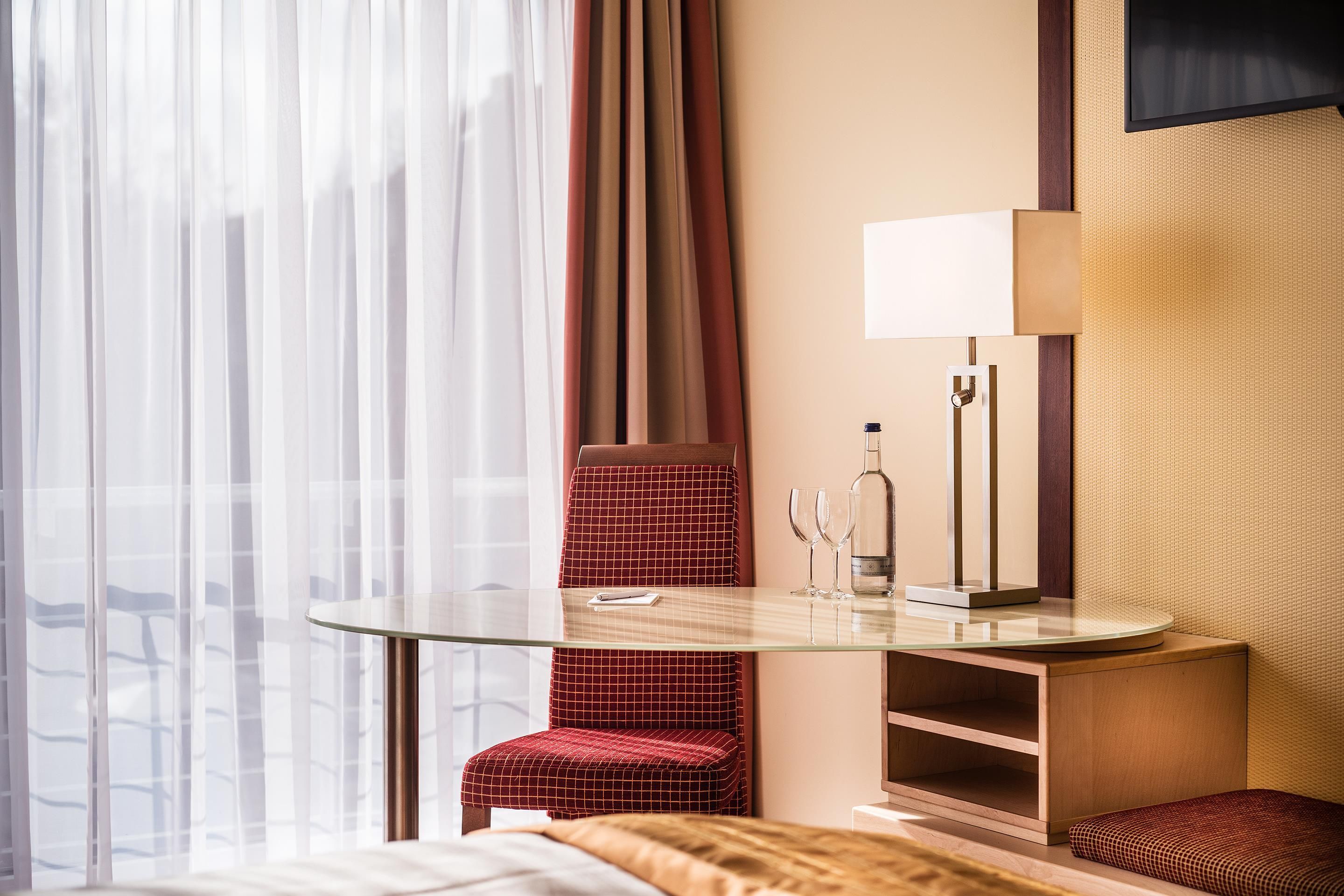 A hotel room with a glass-top desk, a red checkered chair, a bed with a white and brown patterned bedspread, a wall-mounted TV, and a minibar.