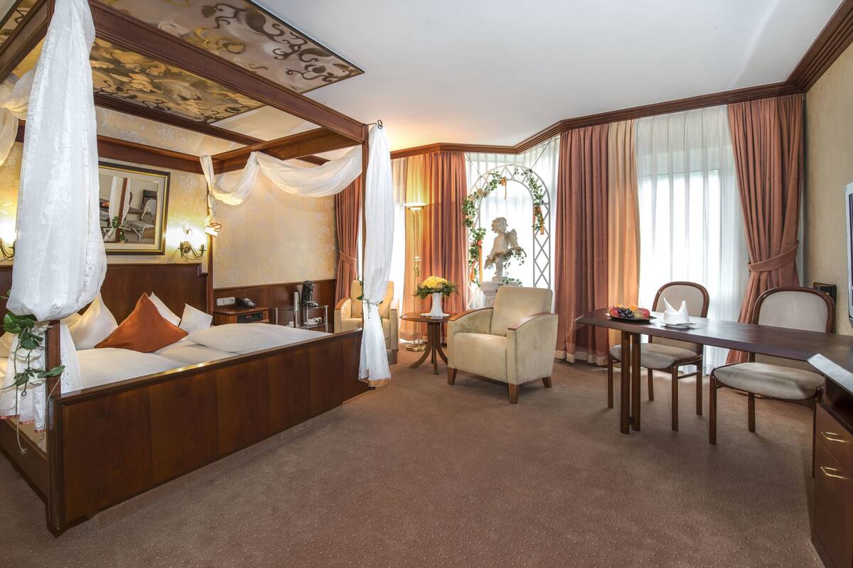 Une chambre d’hôtel avec un lit à baldaquin, un coin salon avec une table et des chaises, une télévision et une plante décorative.