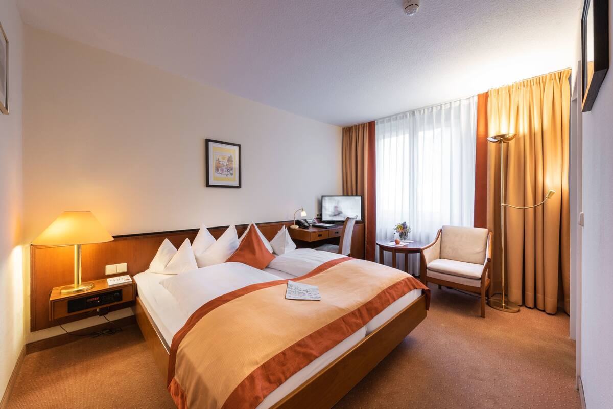 Une chambre d’hôtel avec un grand lit, un bureau avec un ordinateur, une chaise, une lampe et une fenêtre avec des rideaux.