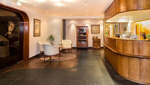 Un espace d’accueil avec un comptoir en bois aux lignes courbes, un coin salon avec deux chaises, une petite table et une plante en pot.