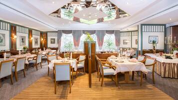 A spacious restaurant with multiple dining tables and chairs, large windows with curtains, and a wooden floor.