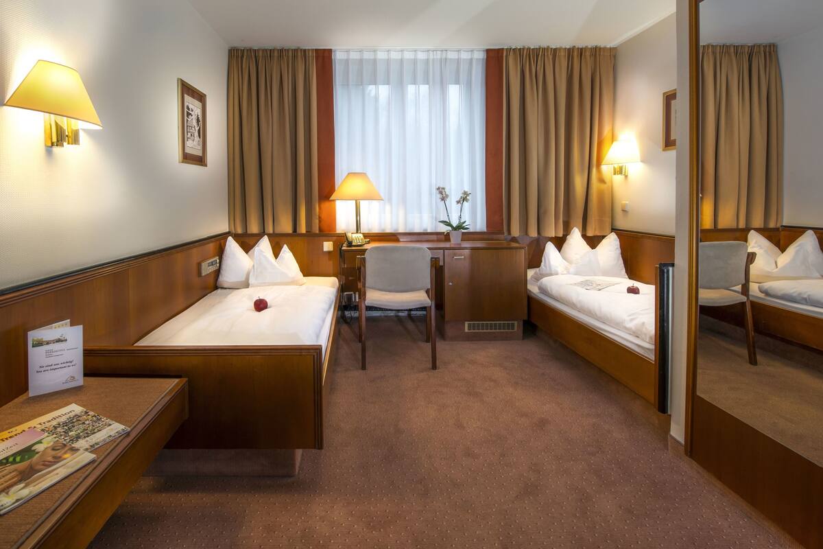 Une chambre d’hôtel avec deux lits simples, un bureau avec une chaise, une petite table sur laquelle se trouve une pomme rouge, et un miroir.
