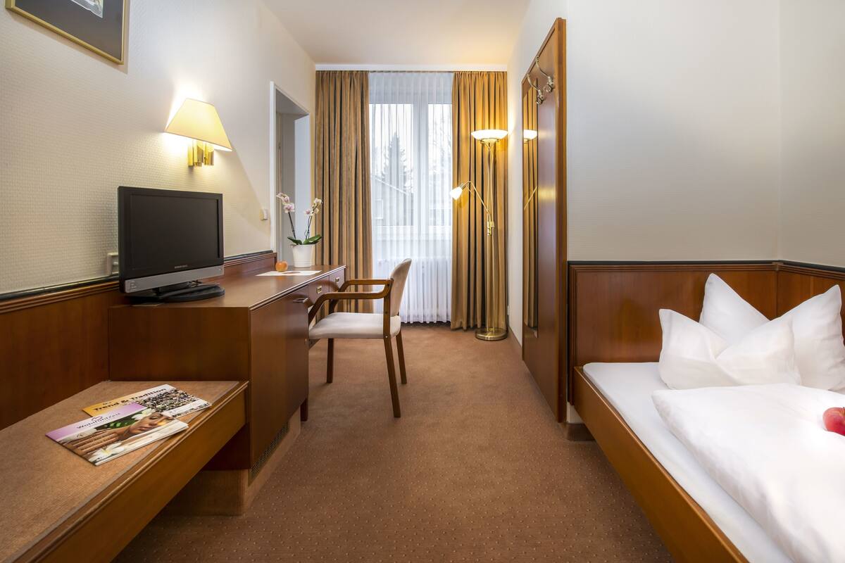 Une chambre d’hôtel avec un lit, un bureau avec un ordinateur, une chaise, une télévision et une fenêtre avec des rideaux.