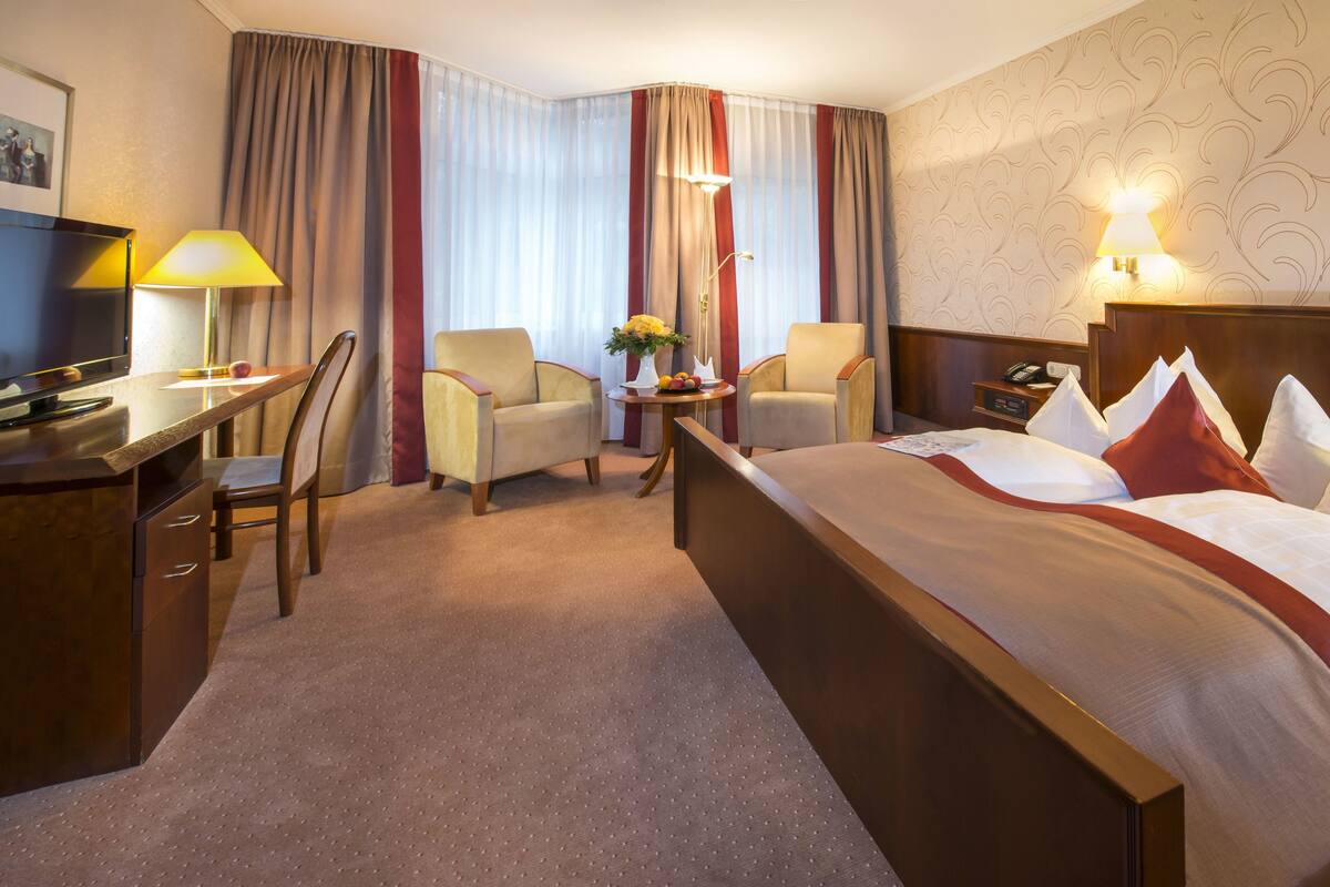 Une chambre d’hôtel comprenant un lit, deux fauteuils, un bureau avec une lampe, une petite table avec un vase de fleurs et un téléphone.