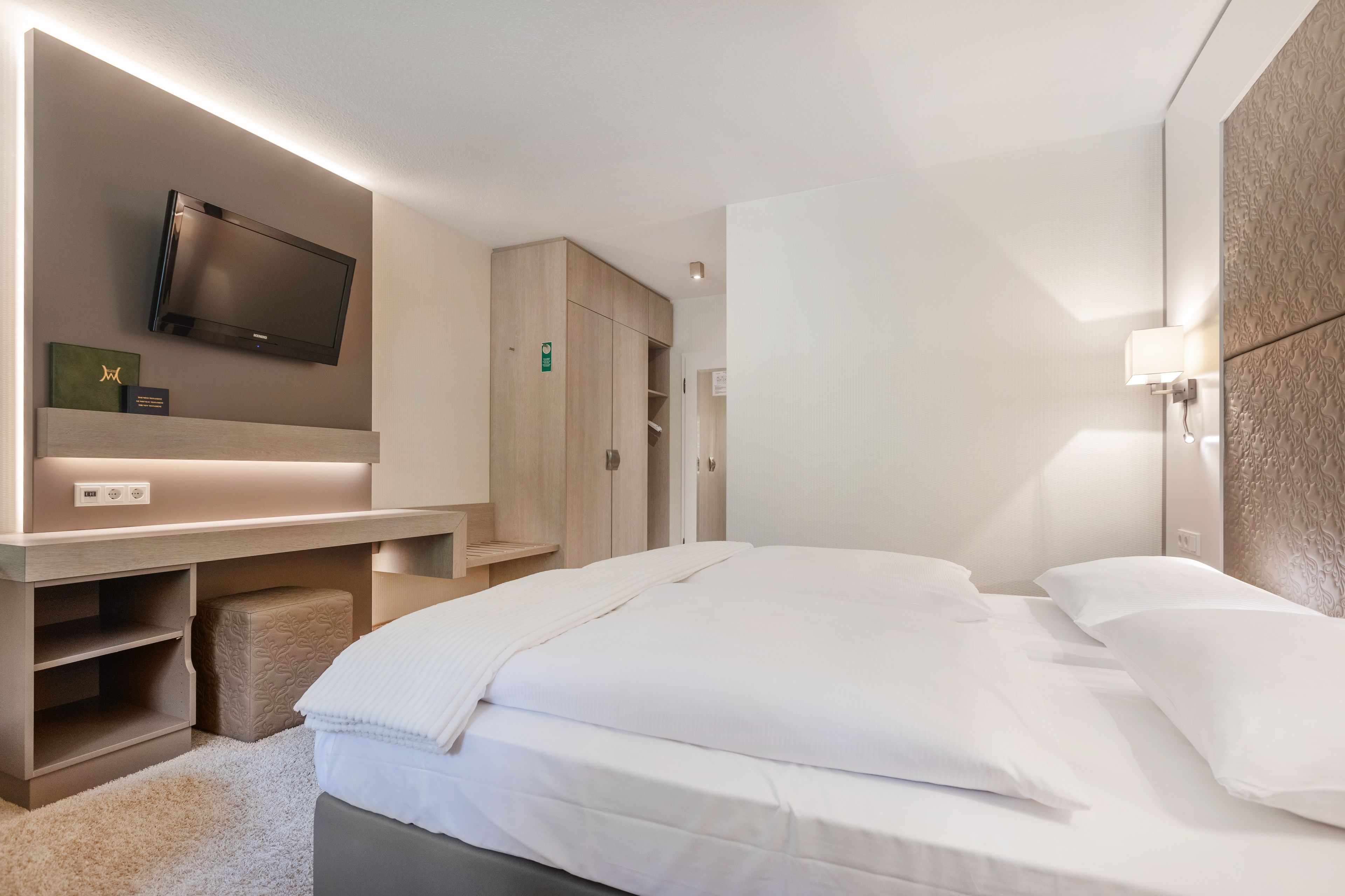A modern hotel room with a bed, a TV mounted on the wall, a wooden wardrobe, and a small desk with a lamp.