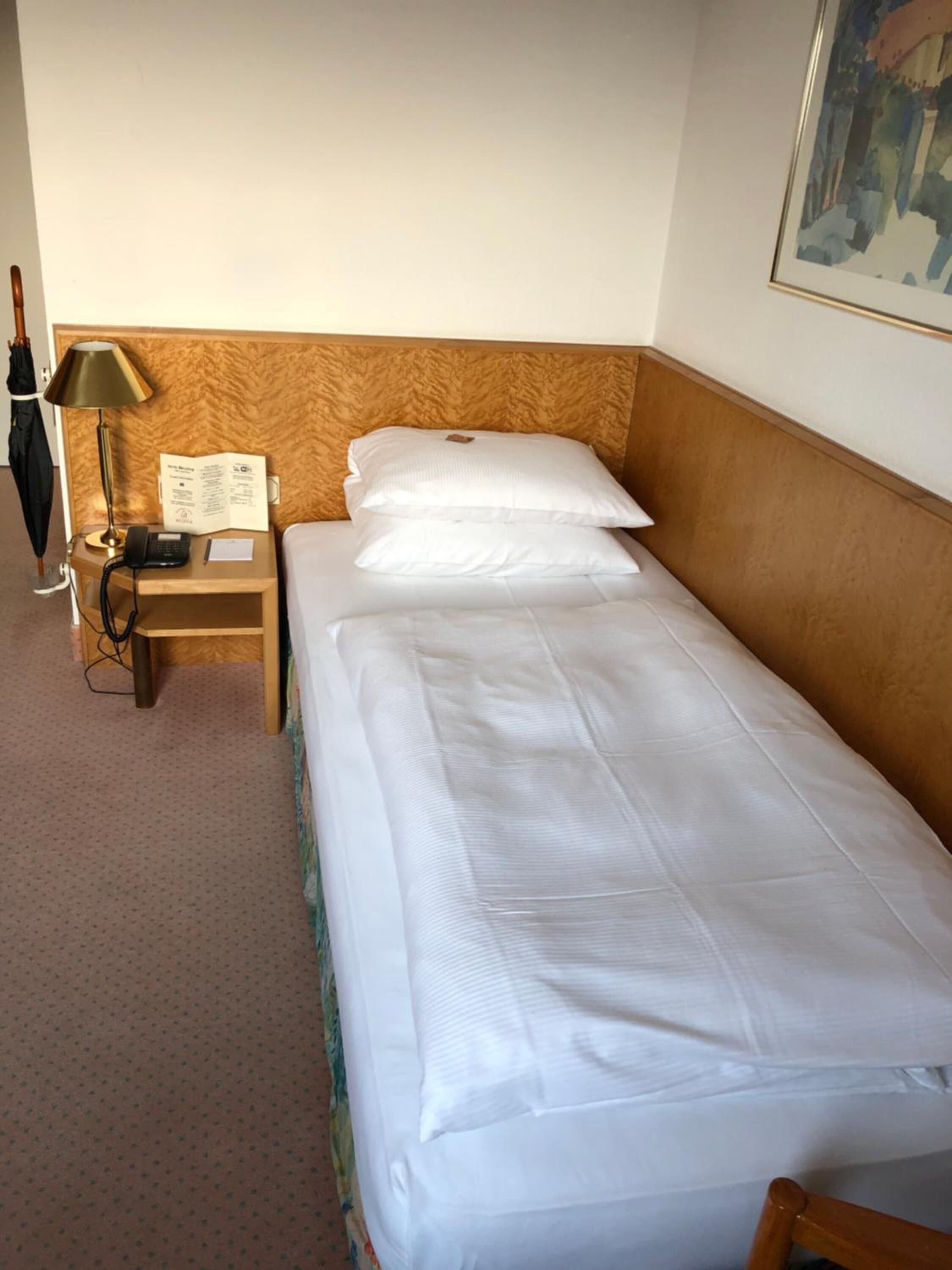 A hotel room with a single bed, a wooden headboard, a nightstand with a lamp, a telephone, and a framed picture on the wall.