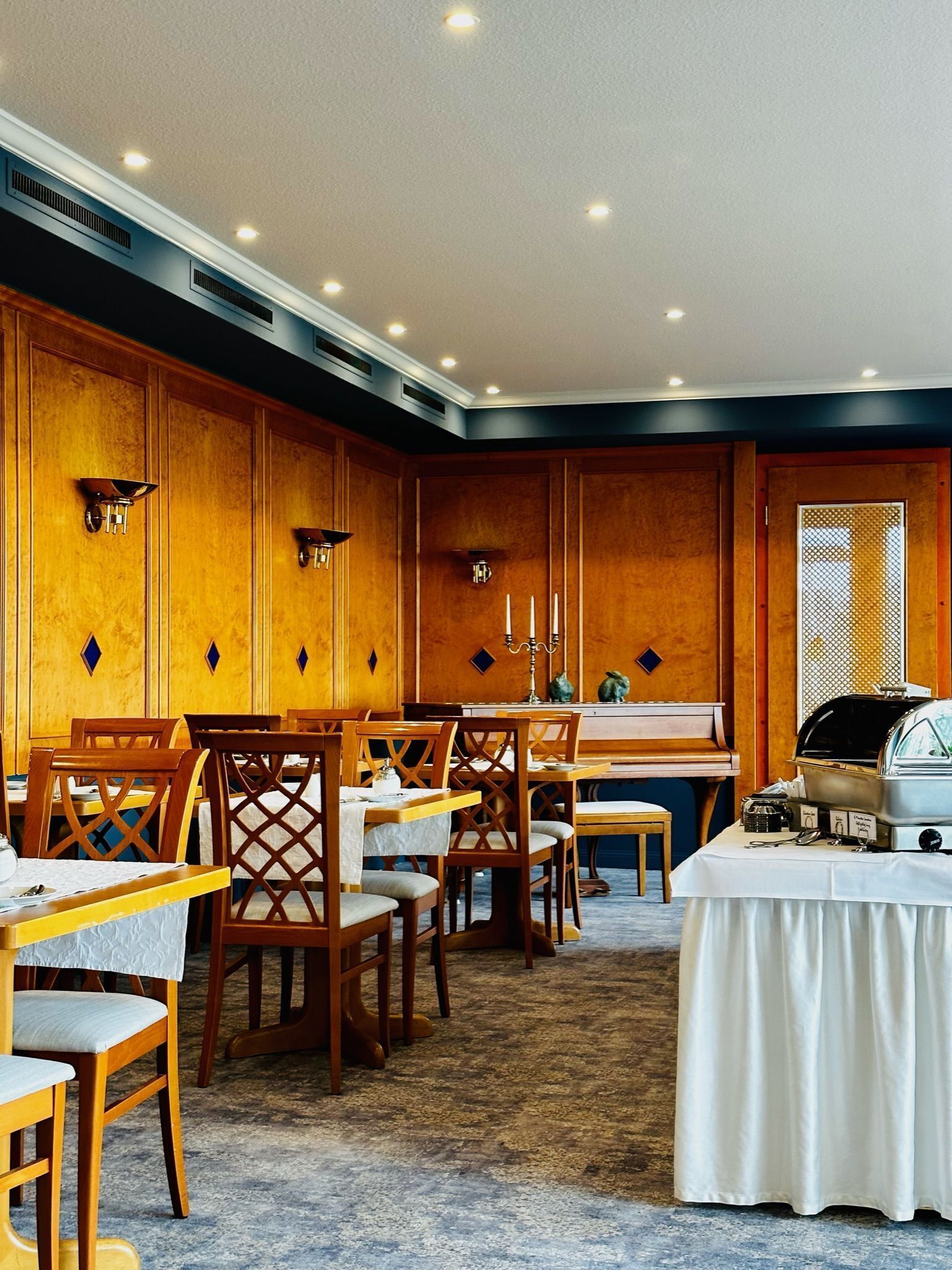 A dining area with wooden paneling, tables, chairs, a piano, and a buffet station.