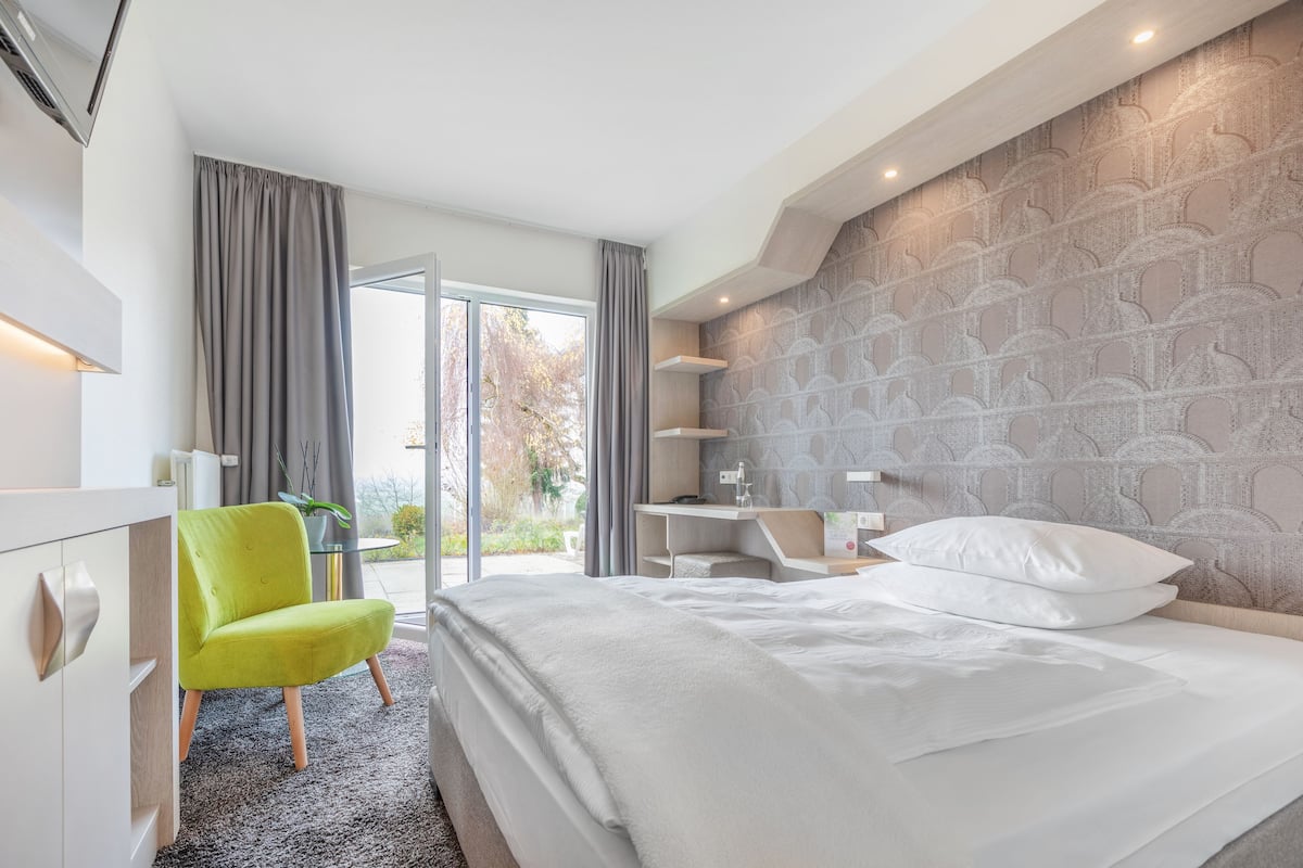 A modern hotel room with a large bed, a bright green chair, a desk, and a view of the outdoors.