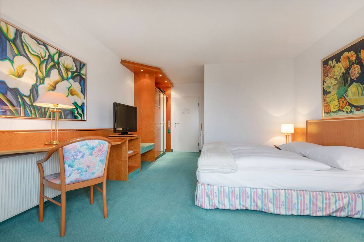 A hotel room with a bed, a desk with a television, a chair, and a painting on the wall.