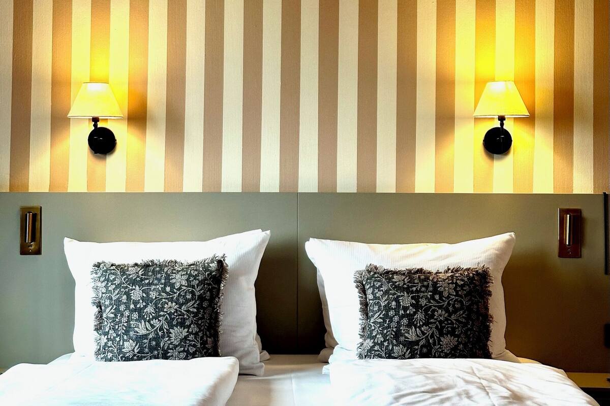 A hotel room with two beds, striped wallpaper, and wall-mounted lamps.