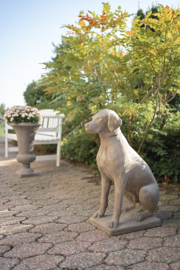 A stone statue of a dog sitting on a paved area with a potted plant and a white chair in the background.