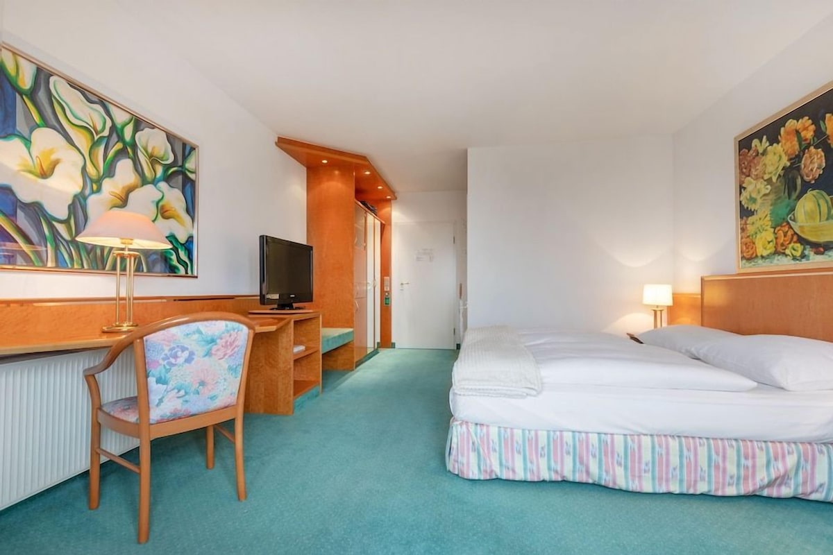 A hotel room with a bed, a desk with a TV, a chair, and two paintings on the walls.