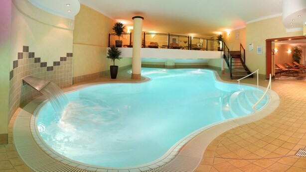 An indoor swimming pool with a curved design, a seating area with lounge chairs, and a staircase leading to an upper level.