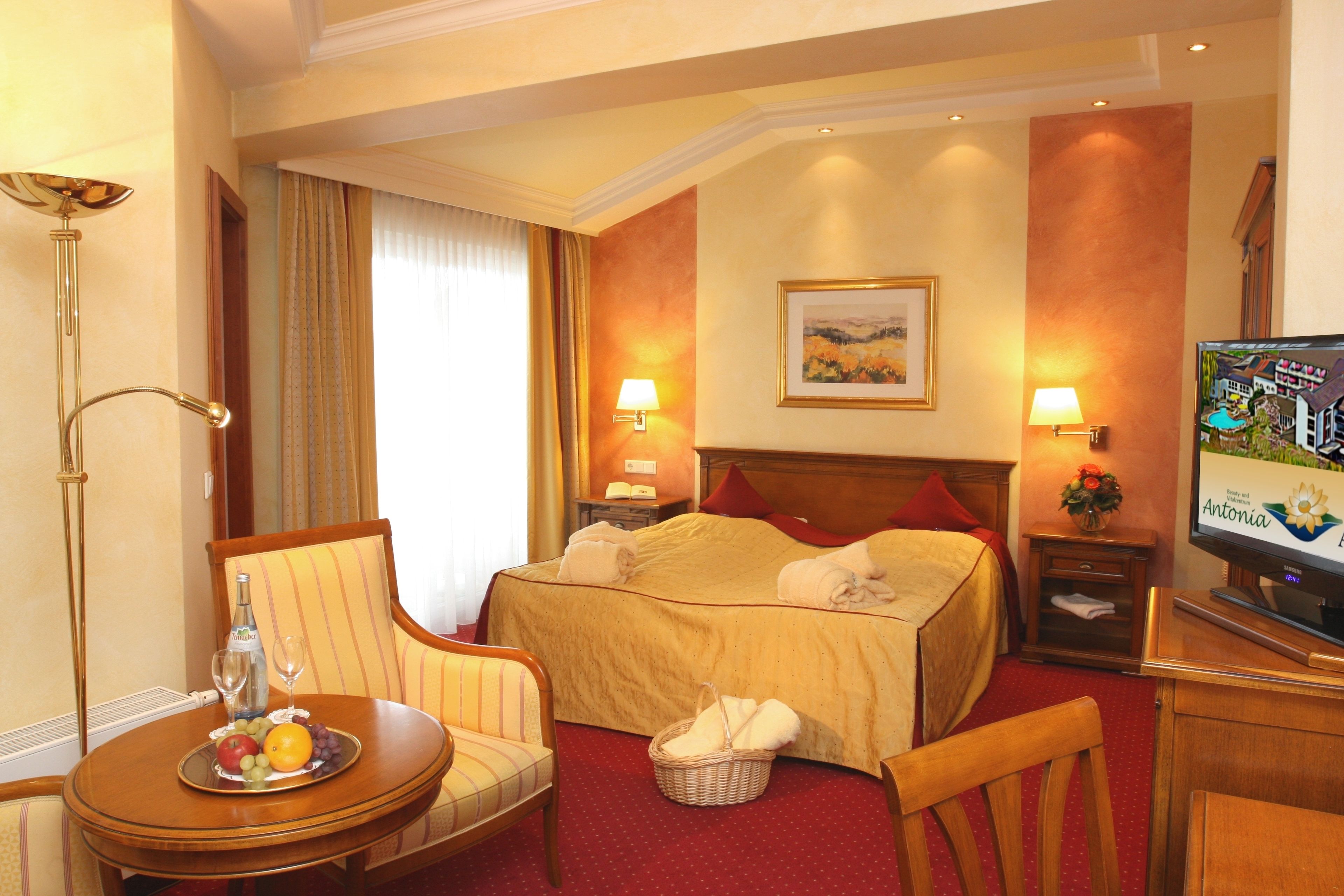 A hotel room with a bed, a television, a dining table with fruit, a chair, a lamp, and a painting on the wall.