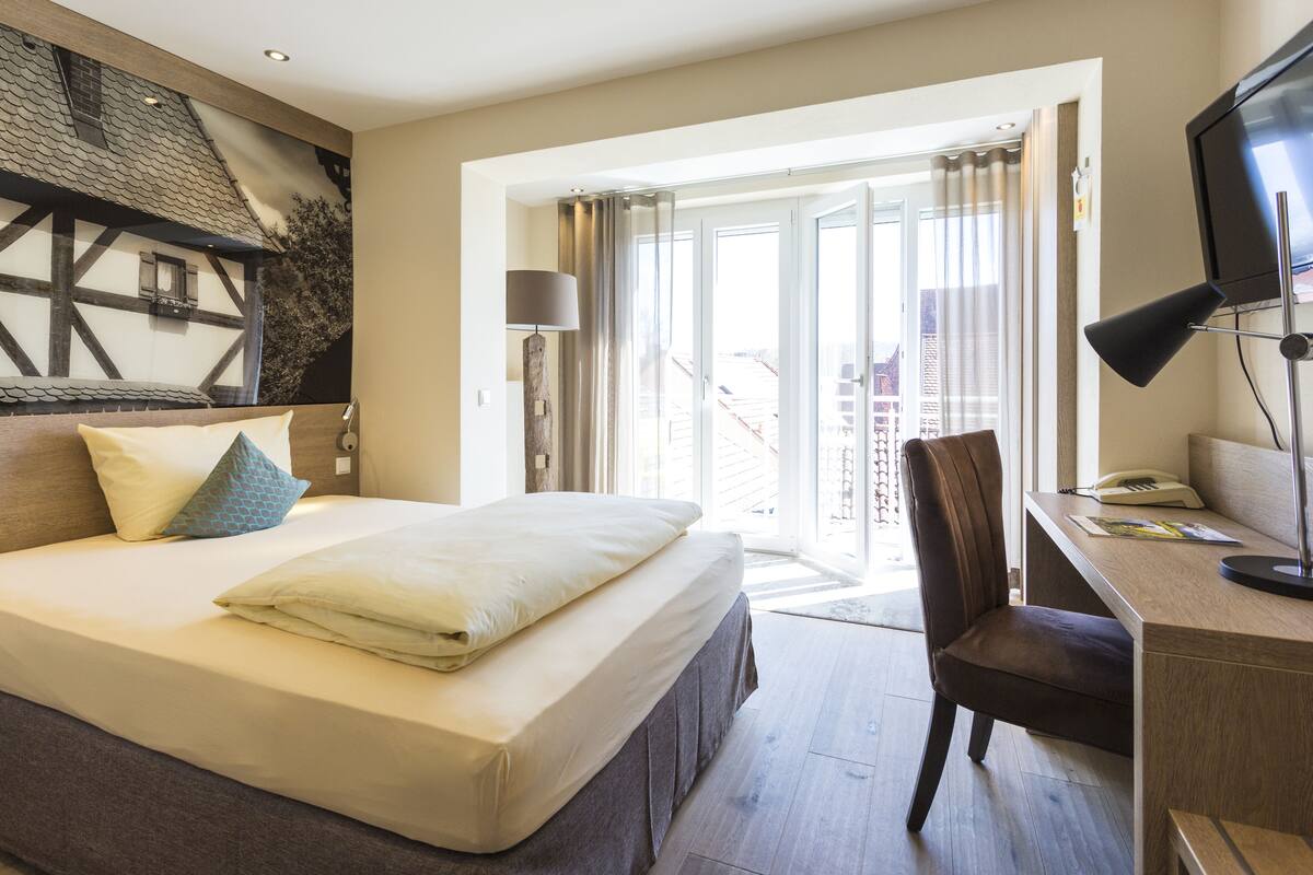 Habitación de hotel con cama, escritorio, silla, televisión y vista a la ciudad por la ventana.