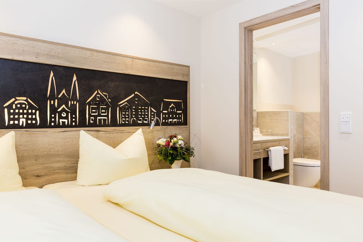 Una habitación de hotel con cama, cabecera de madera, un cuadro con edificios, un florero y un baño visible a través de una puerta abierta.